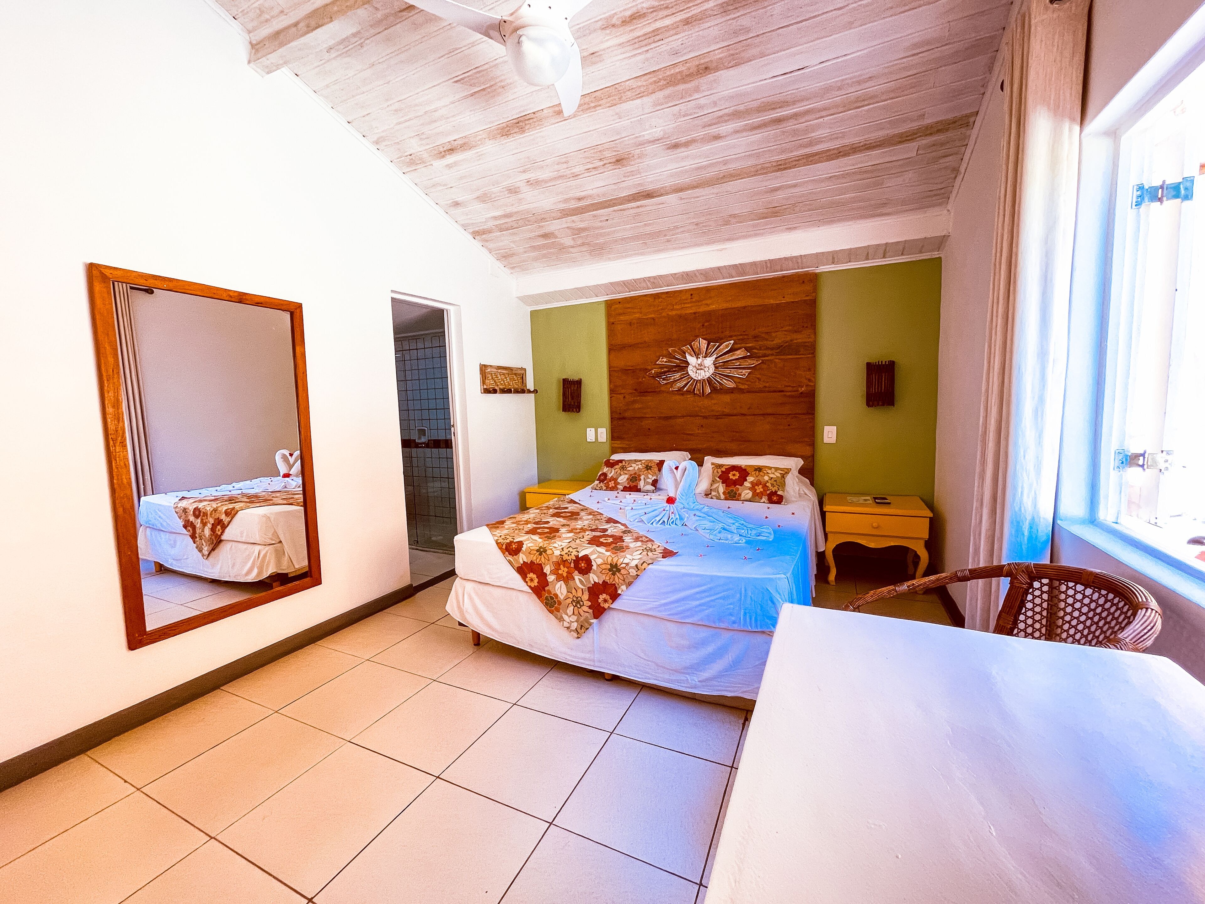 A hotel room with a bed, a chair, a table, a mirror, a wooden headboard, and a window.