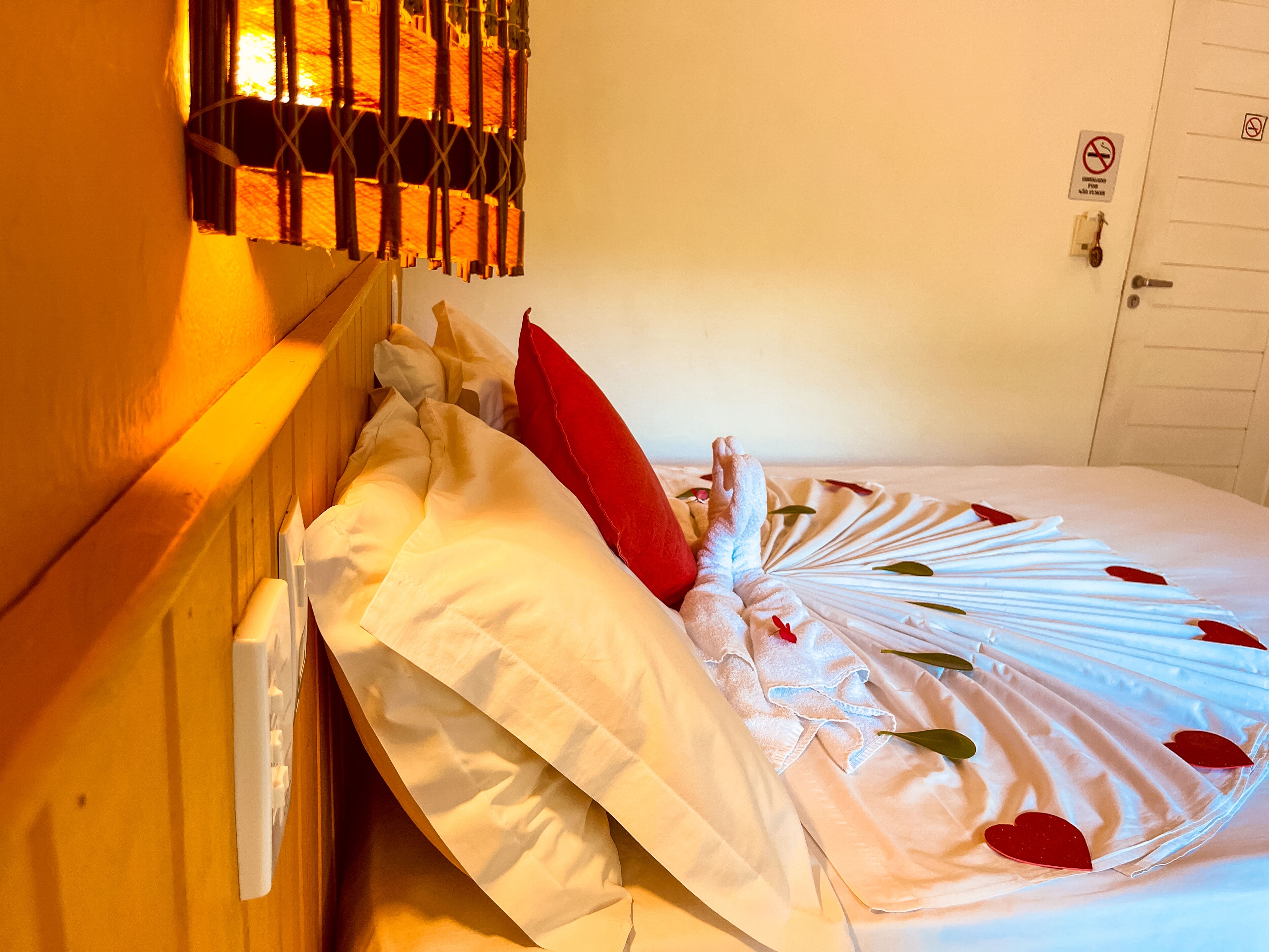 Een hotelkamer met een bed, witte lakens, een rood kussen en een decoratieve ventilator met hartvormige versieringen.