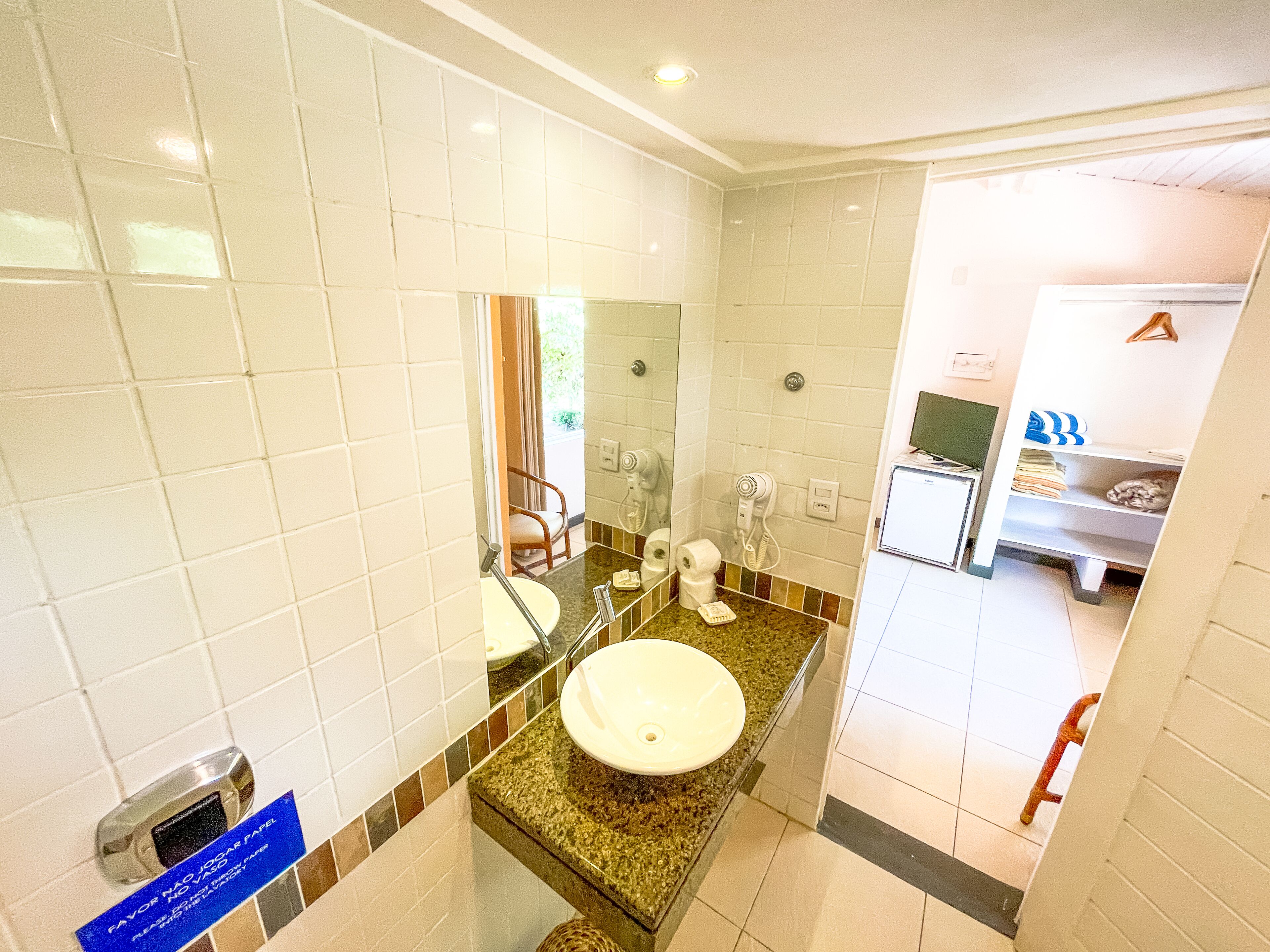 A bathroom with white tiles, a sink, a mirror, a hairdryer, and a shelf with towels.