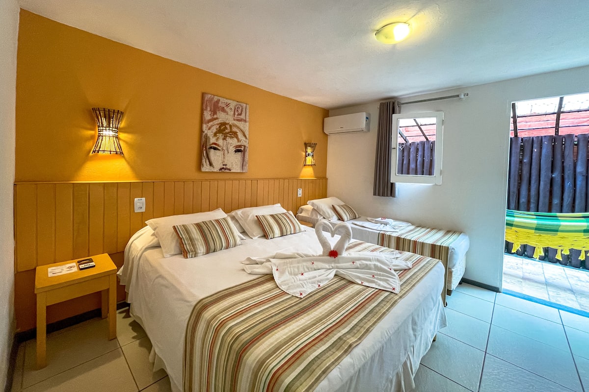 Hotelkamer met twee bedden, een houten hoofdeinde, een wandairconditioner, een raam met gordijnen en een tafeltje met een lamp.