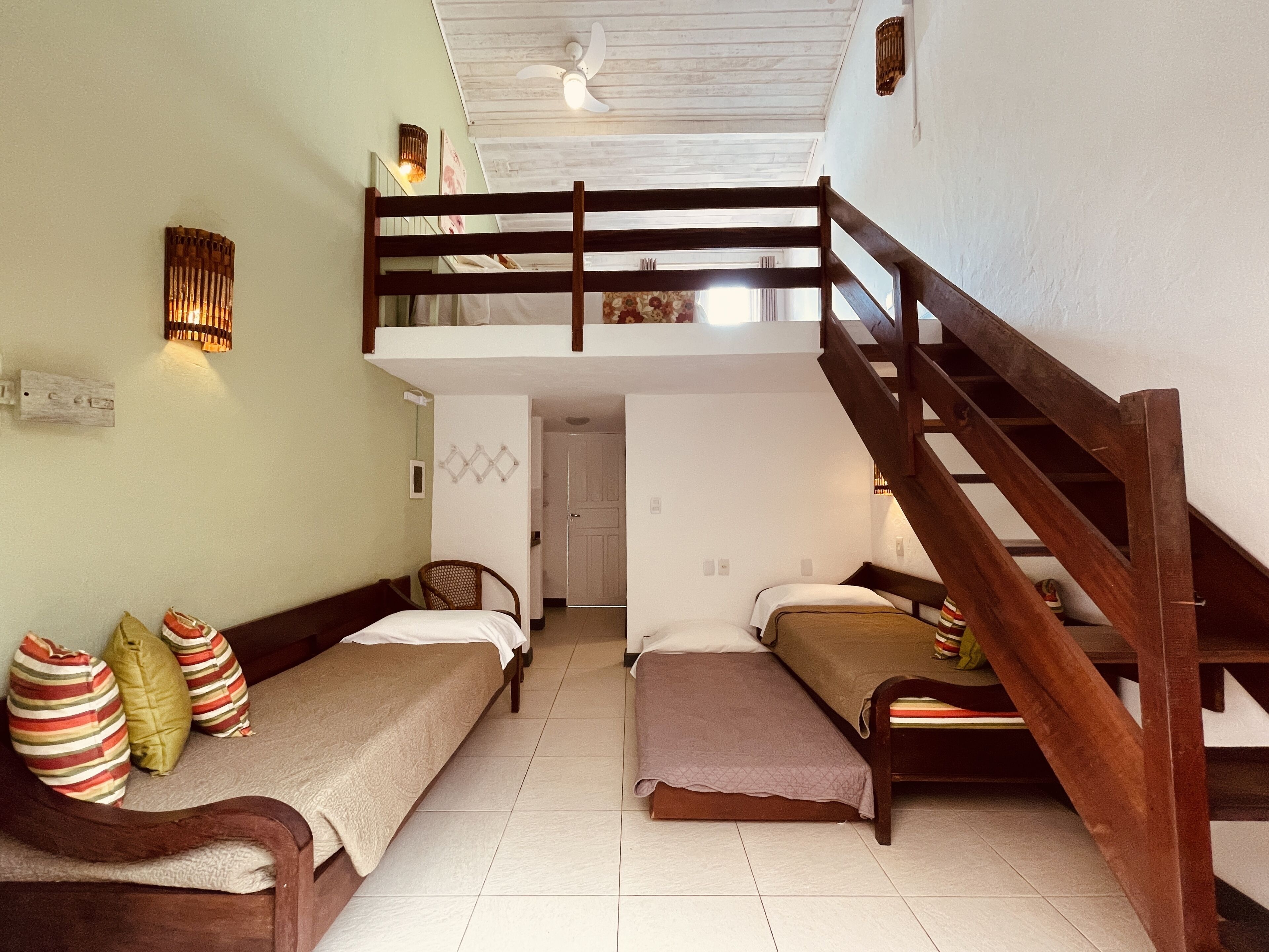 Une chambre de dortoir avec deux lits simples, un lit superposé en bois, un escalier et un ventilateur de plafond.