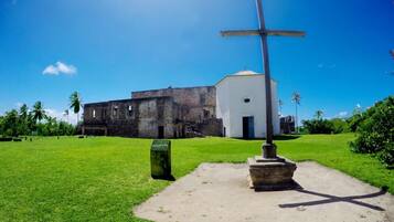 A grassy area with a cross monument, a white building, and a stone structure in the background.