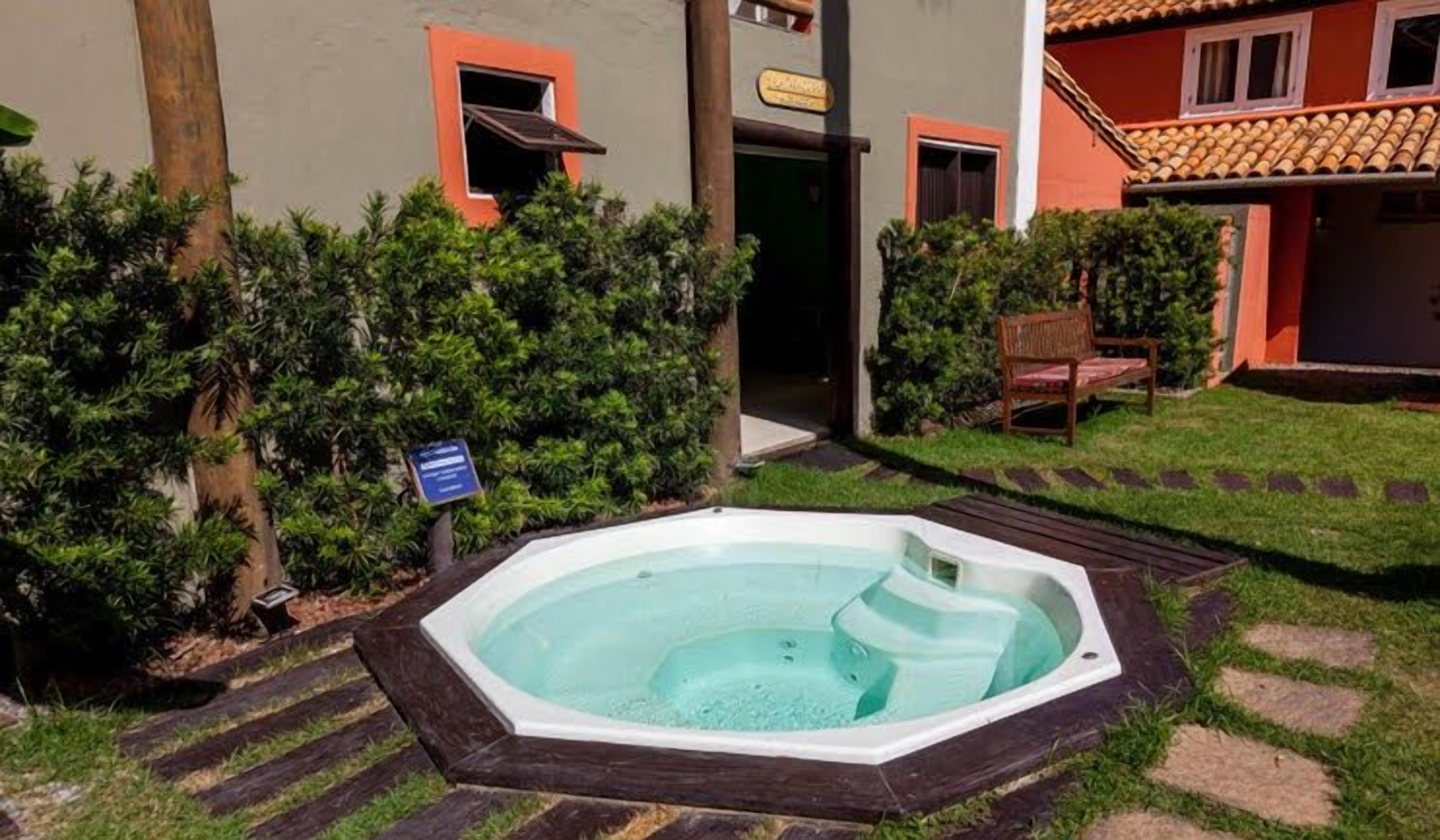 Uma banheira de hidromassagem em um jardim ajardinado com deck de madeira, cercada por vegetação e um edifício com paredes laranja e cinzas.