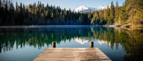 Un muelle de madera se adentra en un lago tranquilo, con una orilla boscosa y una montaña al fondo.