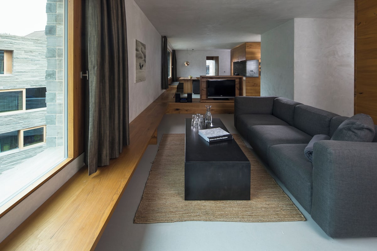 A modern living room with a sofa, a coffee table, and a TV. There is a window with curtains, a rug, and a view of the outside.