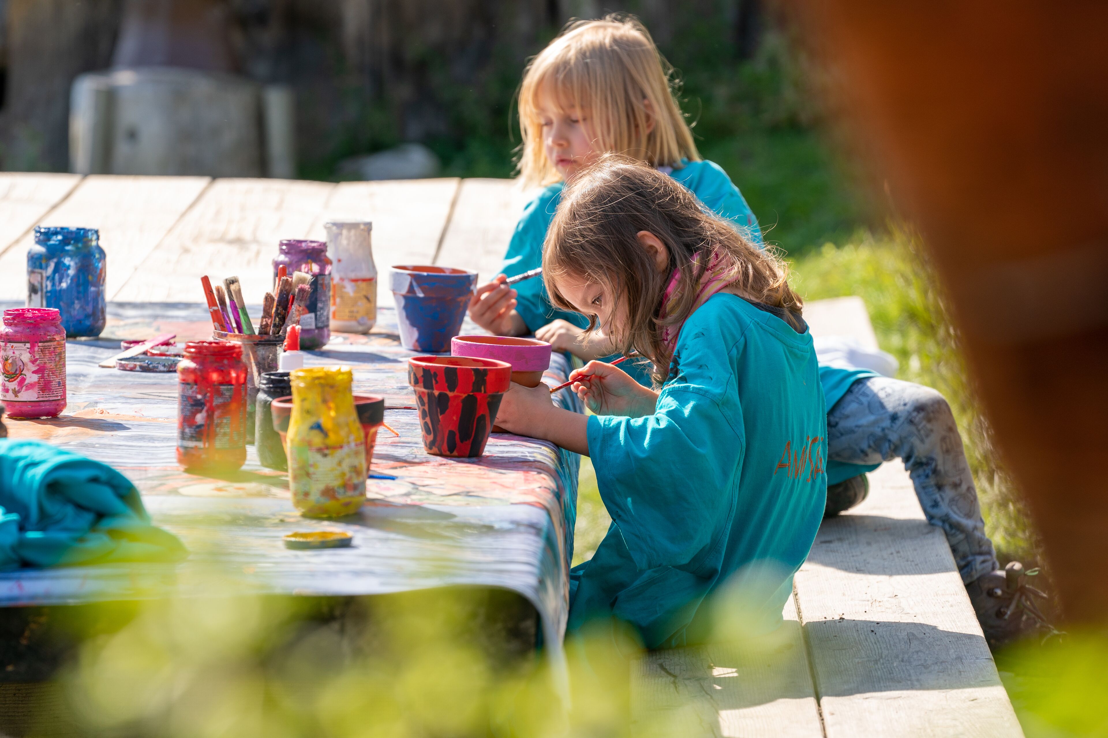 Two children painting outdoors on a wooden table with various paint containers and brushes.