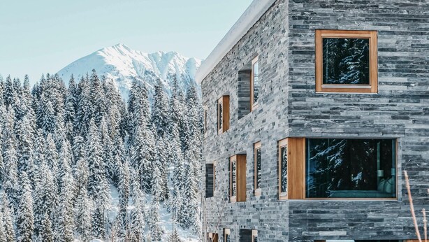 A modern building with a stone facade and large windows, surrounded by snow-covered trees and mountains.
