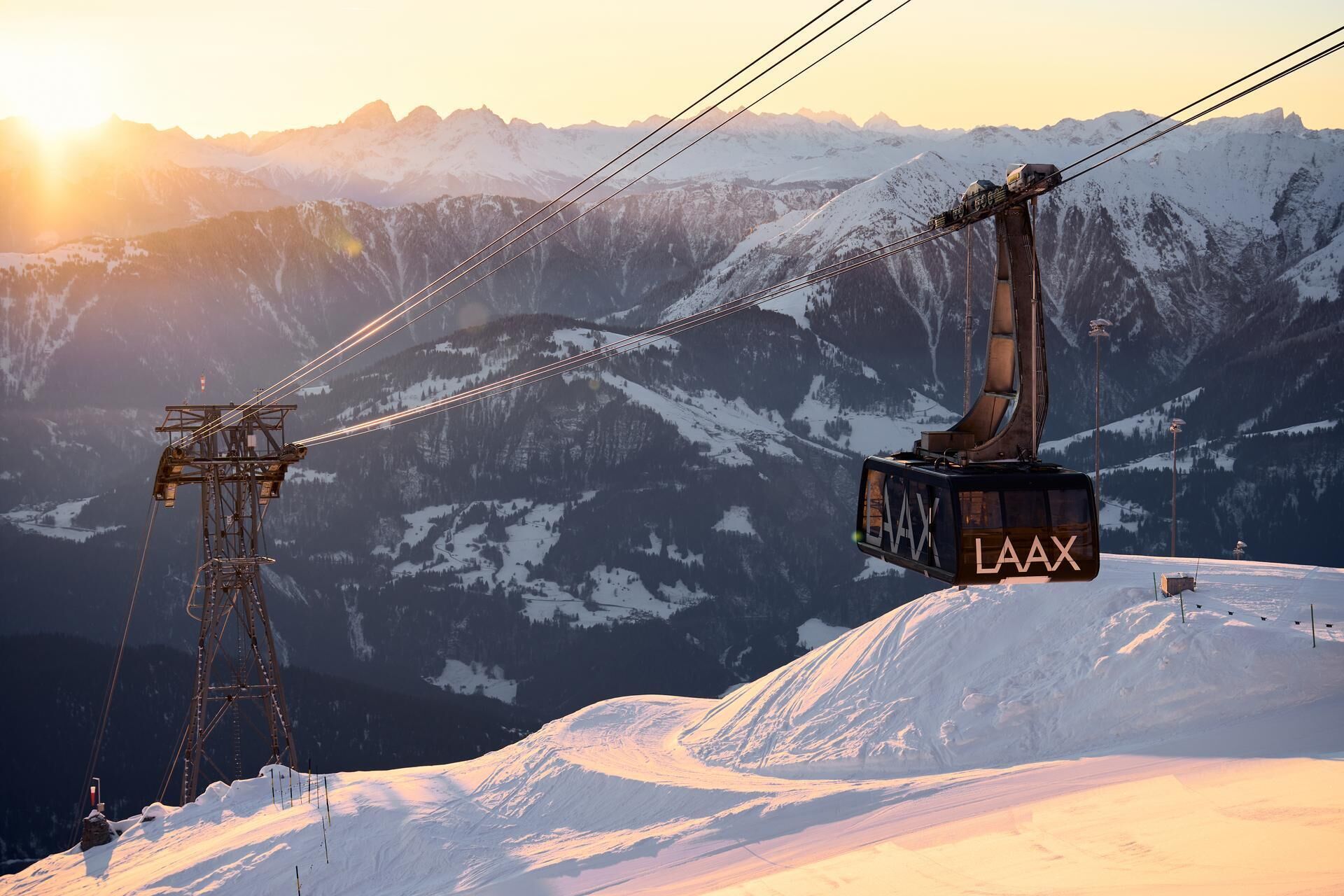 En gondol med "LAAX" på, hængt op i kabler med et bjergrigt landskab i baggrunden under solnedgang.