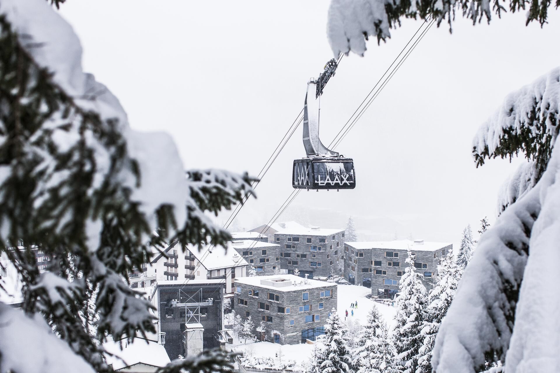 Un telesilla con cabinas suspendidas por cables, rodeado de edificios y árboles cubiertos de nieve.