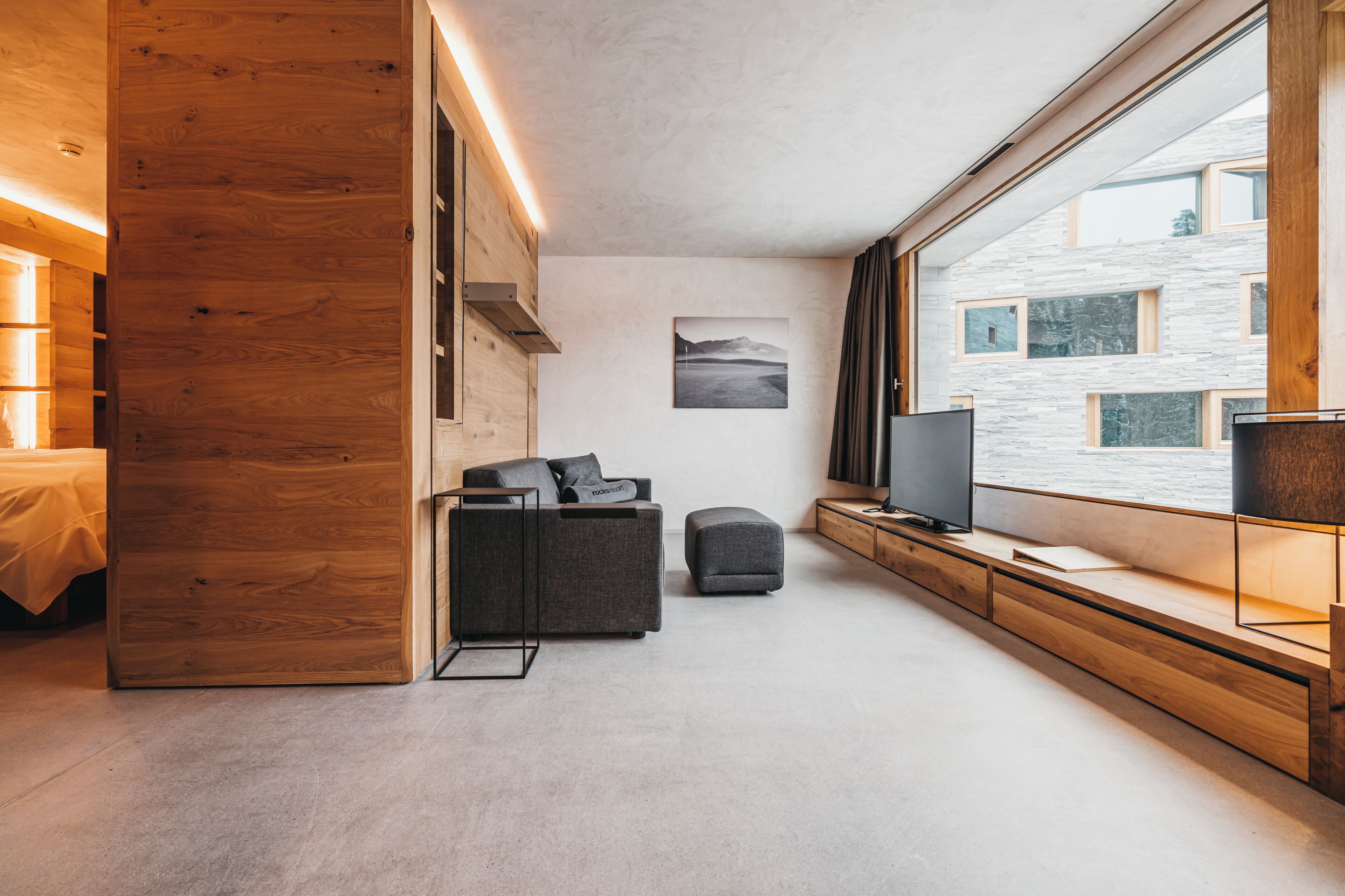 Un salon moderne avec un mur d’accent en bois, un canapé, un petit pouf, un téléviseur et une fenêtre donnant sur un immeuble.