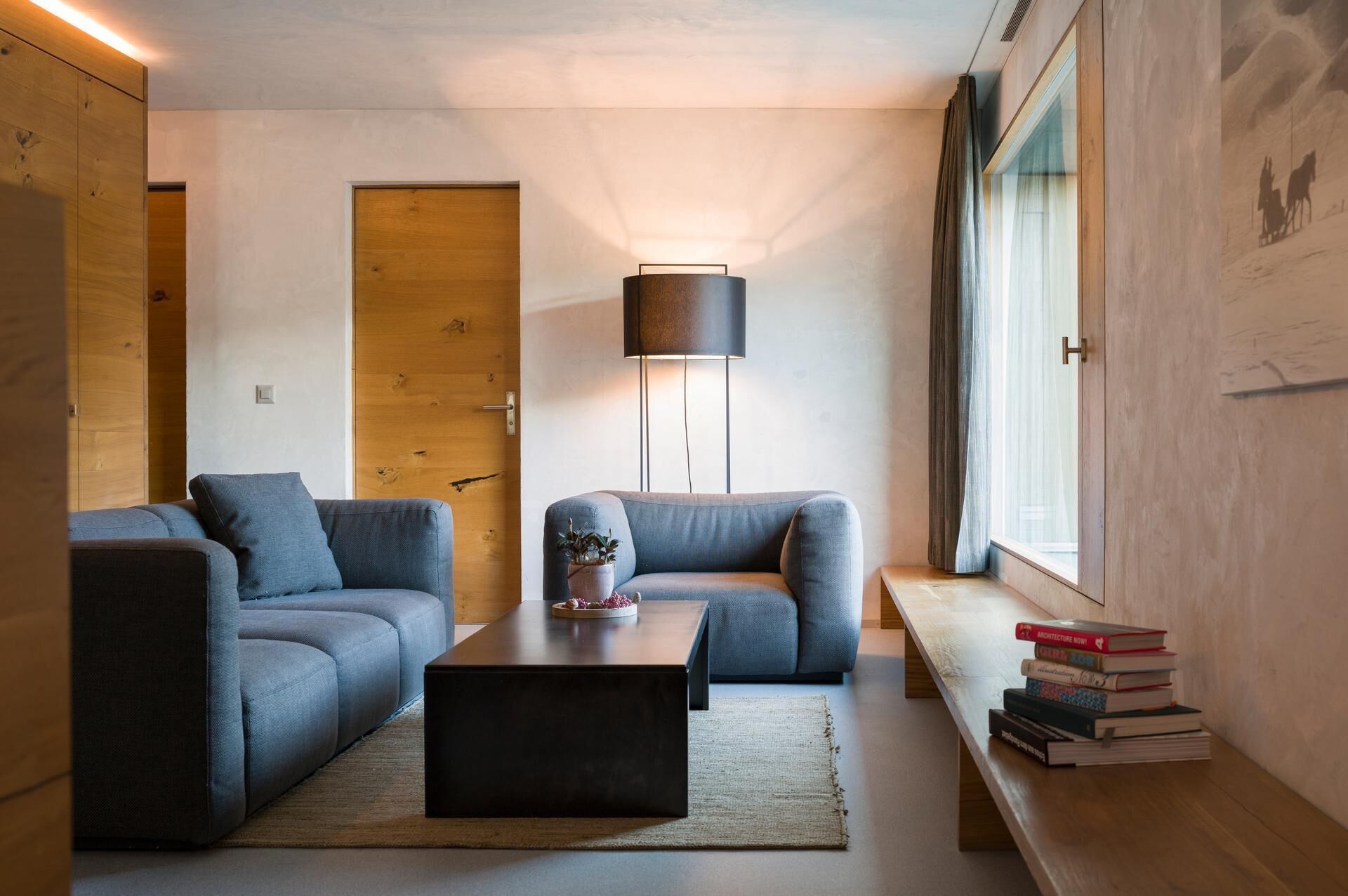 A modern living room with a sofa, a coffee table, and a lamp. There is a wooden door and a window with curtains.