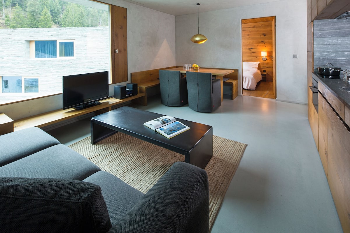 A modern living room with a sofa, a coffee table, and a TV. There is a wooden dining table with chairs and a kitchen area with a sink and stove.