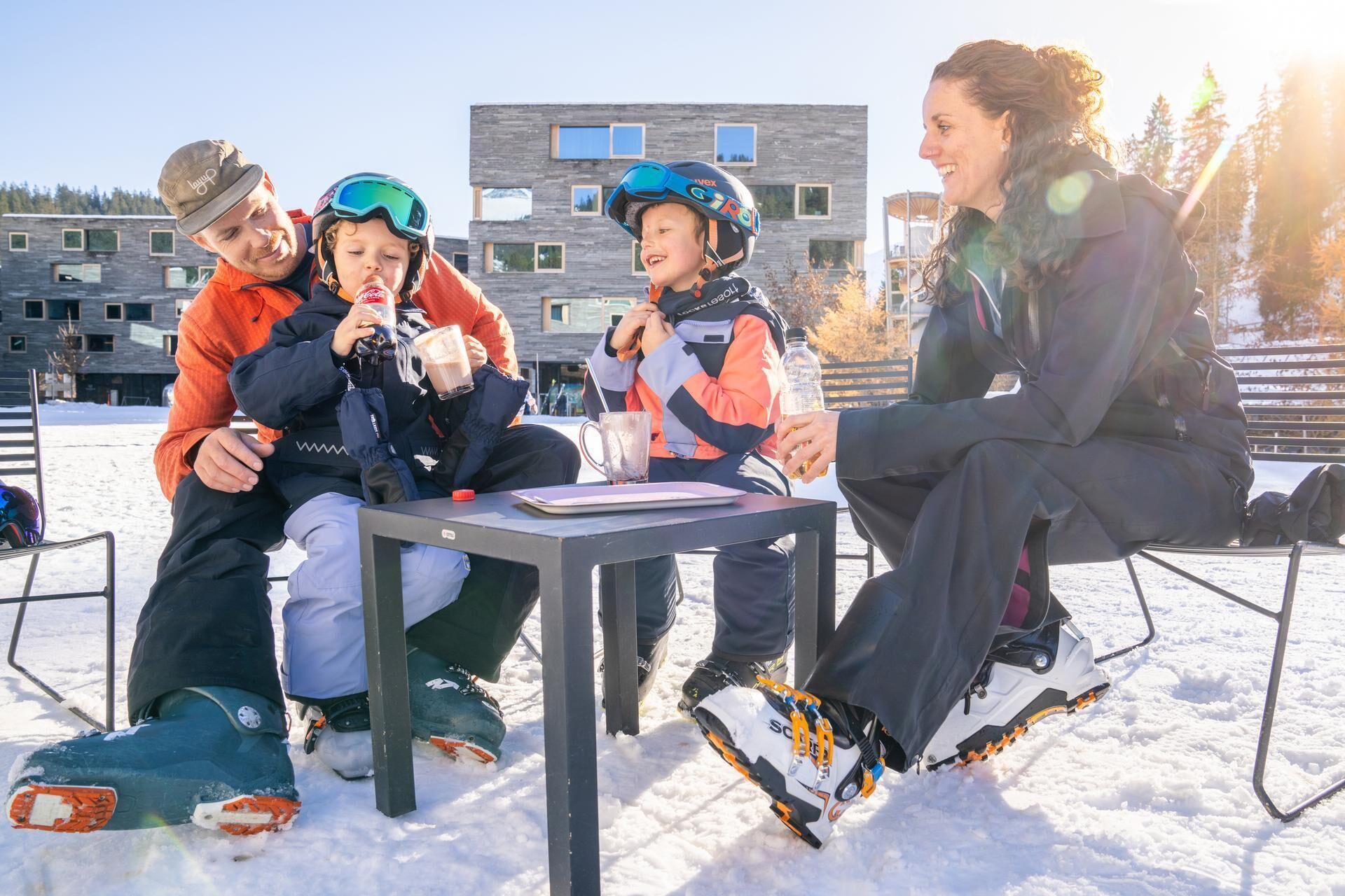 A group of people sitting around a table outdoors in a snowy environment, with a building in the background.
