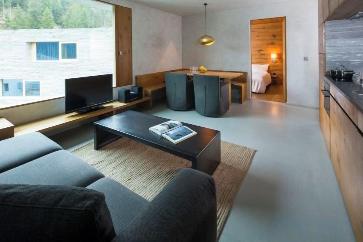Ein modernes Wohnzimmer mit einer Couch, einem Couchtisch und einem Fernseher. Es gibt einen Küchenbereich mit Schränken und einem Esstisch.