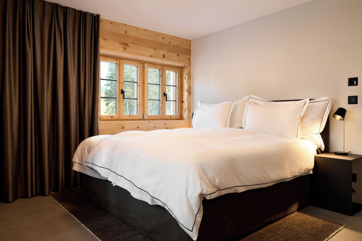 A bedroom with a large bed, wooden walls, a window, and a nightstand with a lamp.