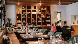 A restaurant interior with multiple dining tables set with glasses and cutlery, a wine rack in the background, and a cozy seating area with cushions.