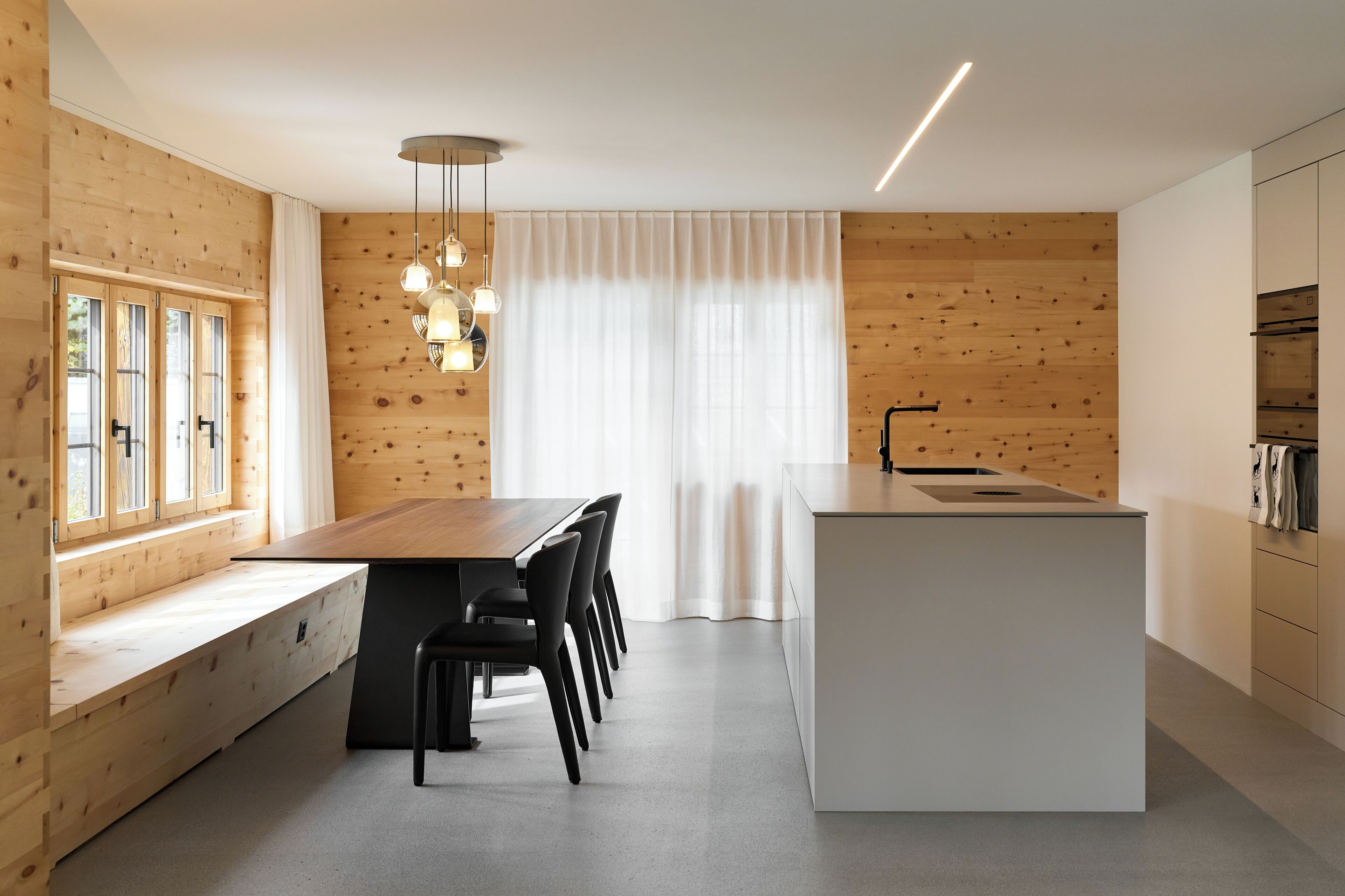 A modern kitchen with a wooden wall, a dining area with a long table and chairs, and a large window with curtains.