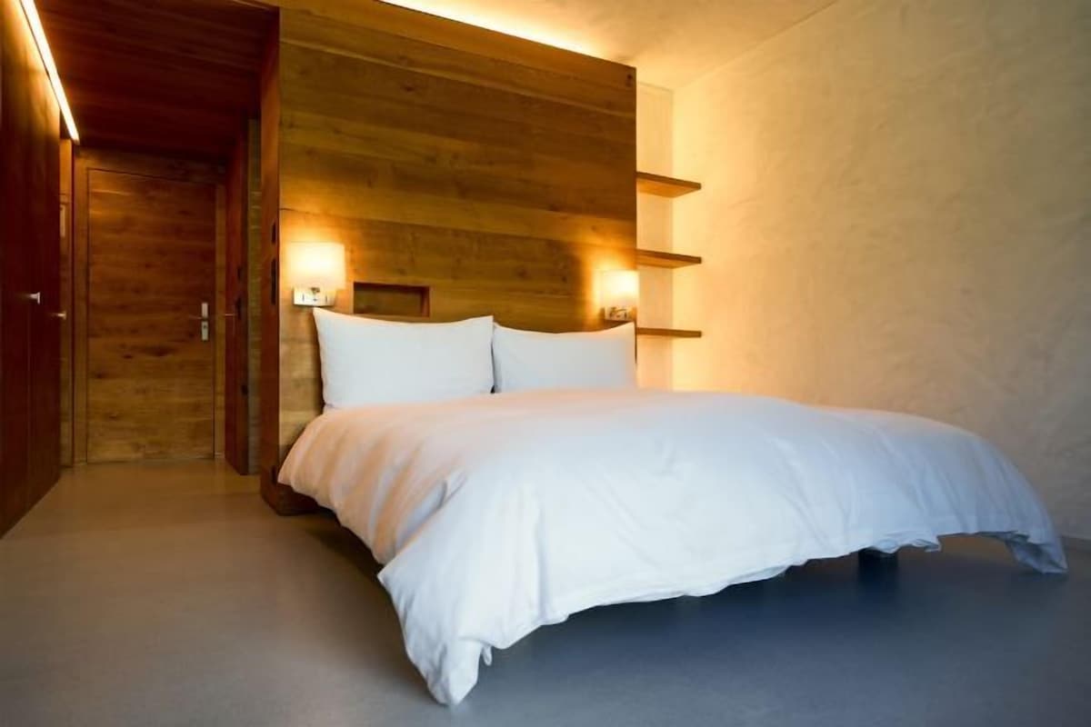 Quarto com cabeceira de madeira, cama com roupa de cama branca e luminárias fixadas na parede.