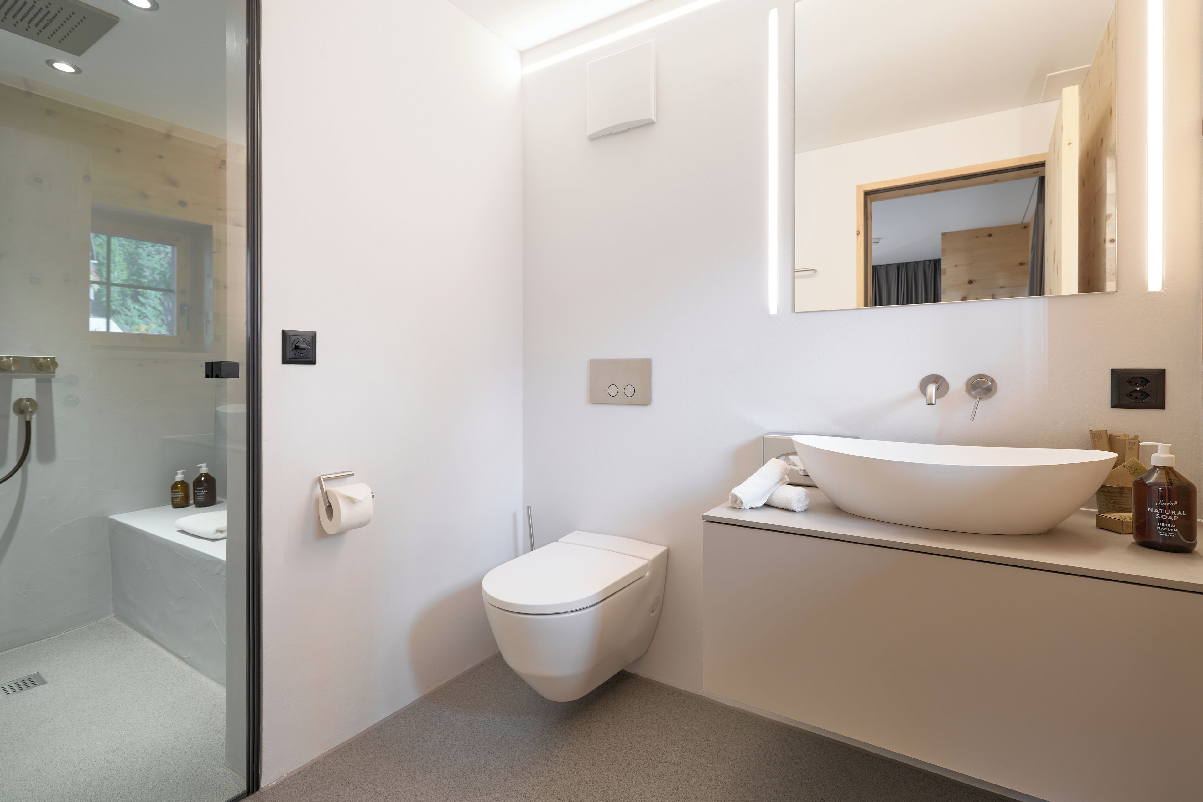 Ein modernes Badezimmer mit Duschkabine aus Glas, Toilette in Weiß, großem Waschbecken mit Haube und einem Spiegel mit integrierter Beleuchtung.