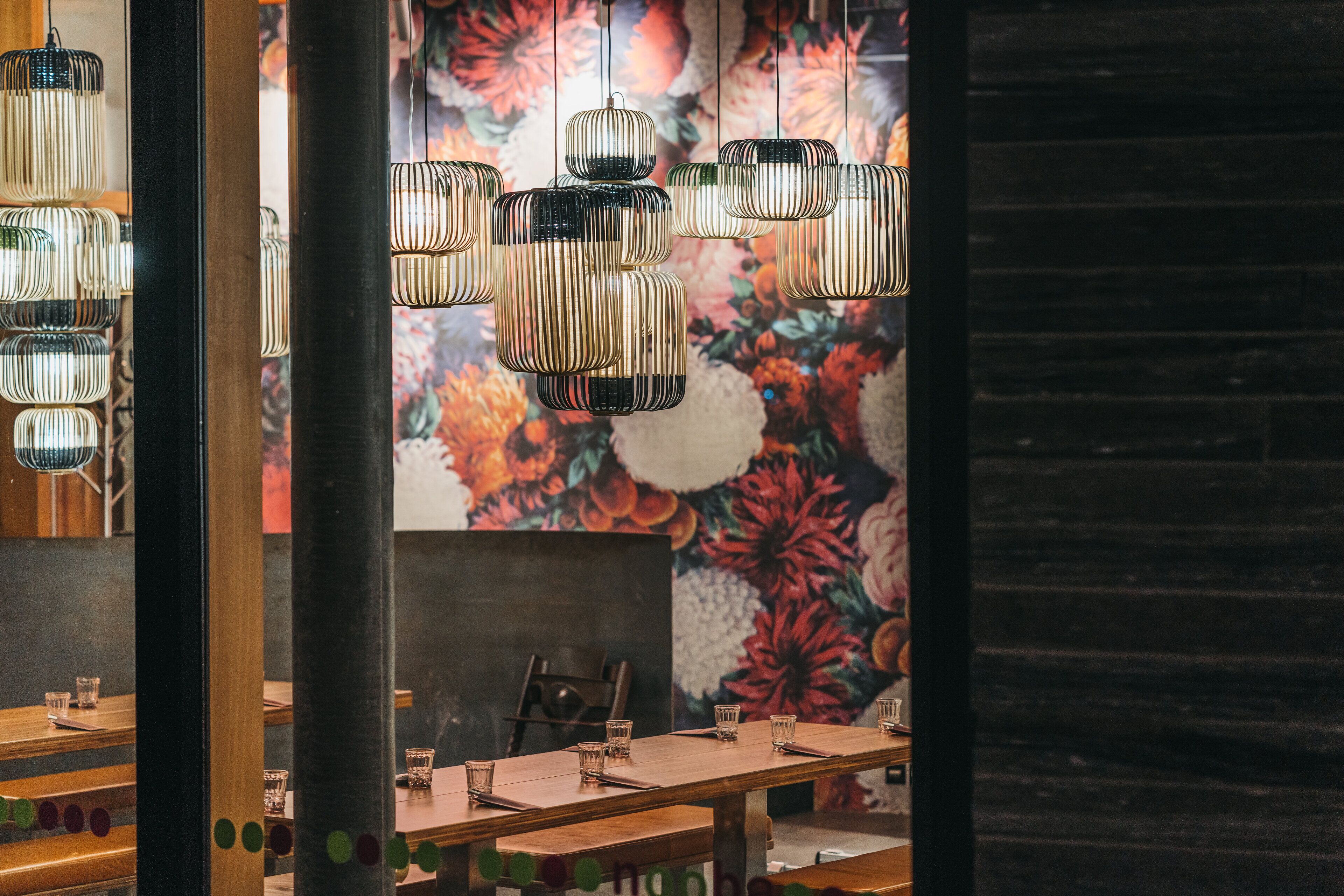 A restaurant interior with a long wooden table, hanging cylindrical light fixtures, and a floral mural on the wall.