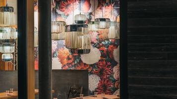 A restaurant interior with a long wooden table, hanging cylindrical light fixtures, and a floral mural on the wall.