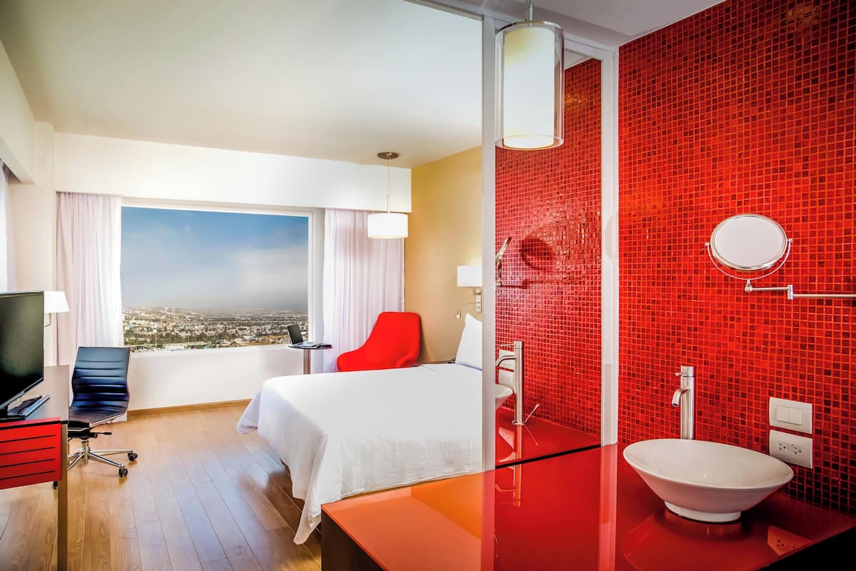 A hotel room with a bed, a desk, a chair, a TV, a window with a view, a bathroom with a red tiled wall, a sink, and a mirror.