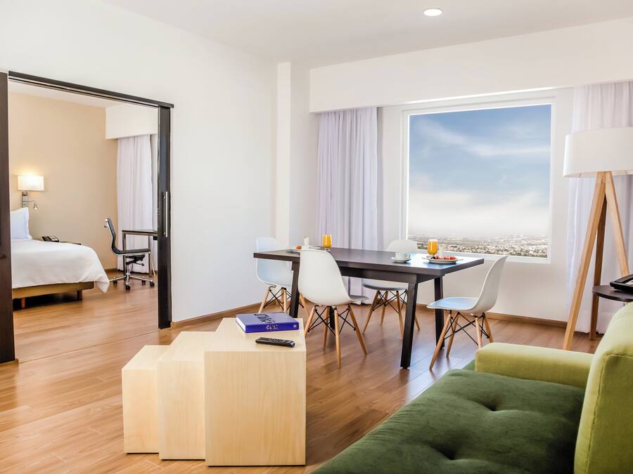 A hotel room with a bed, a desk with a chair, a green sofa, a TV, and a window with a view.