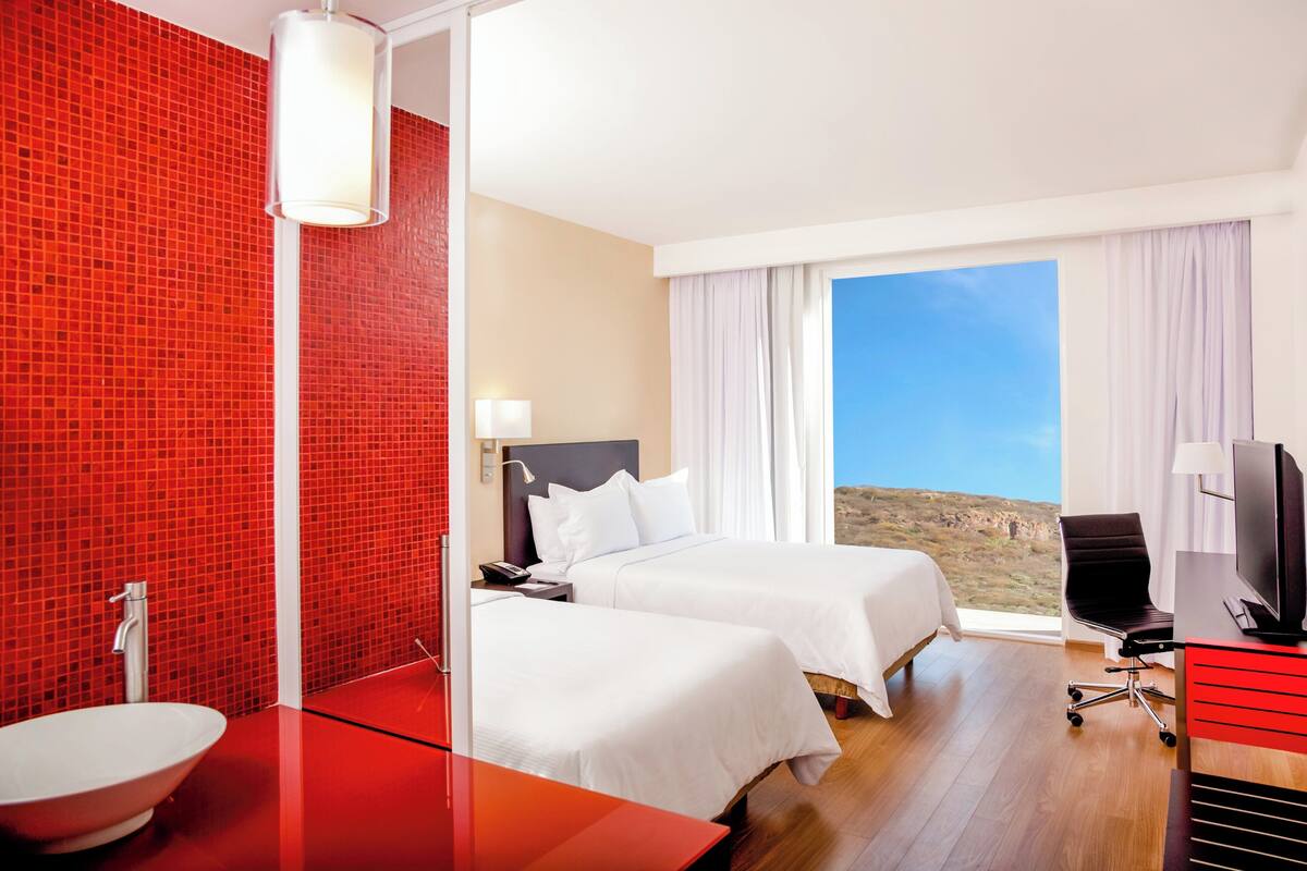 A hotel room with two beds, a red-tiled wall, a large window with a view, a TV, and a desk.