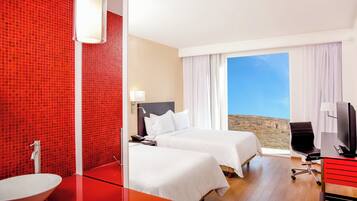 Habitación de hotel con dos camas, pared de baldosas rojas, un ventanal con vistas, televisor y escritorio.