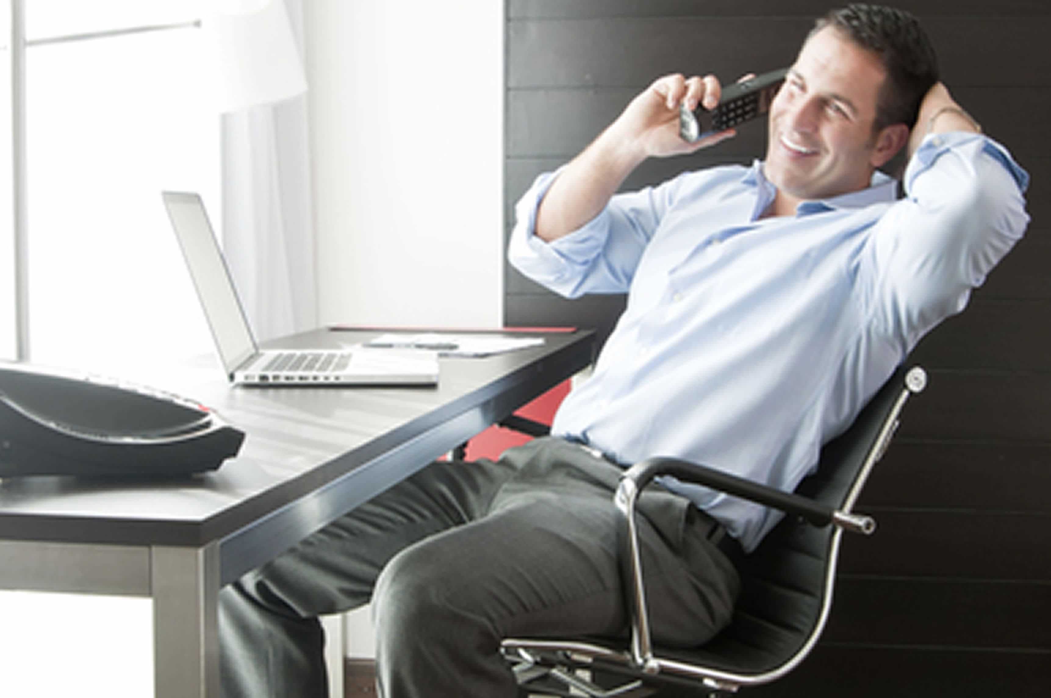 A man sitting in a modern office chair at a desk with a laptop, a smartphone, and a briefcase.