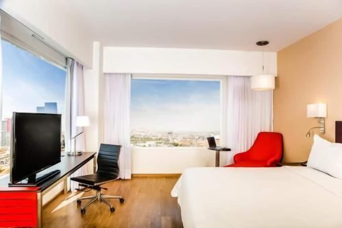 A hotel room with a bed, a desk with a TV, a red chair, and a view of the city.