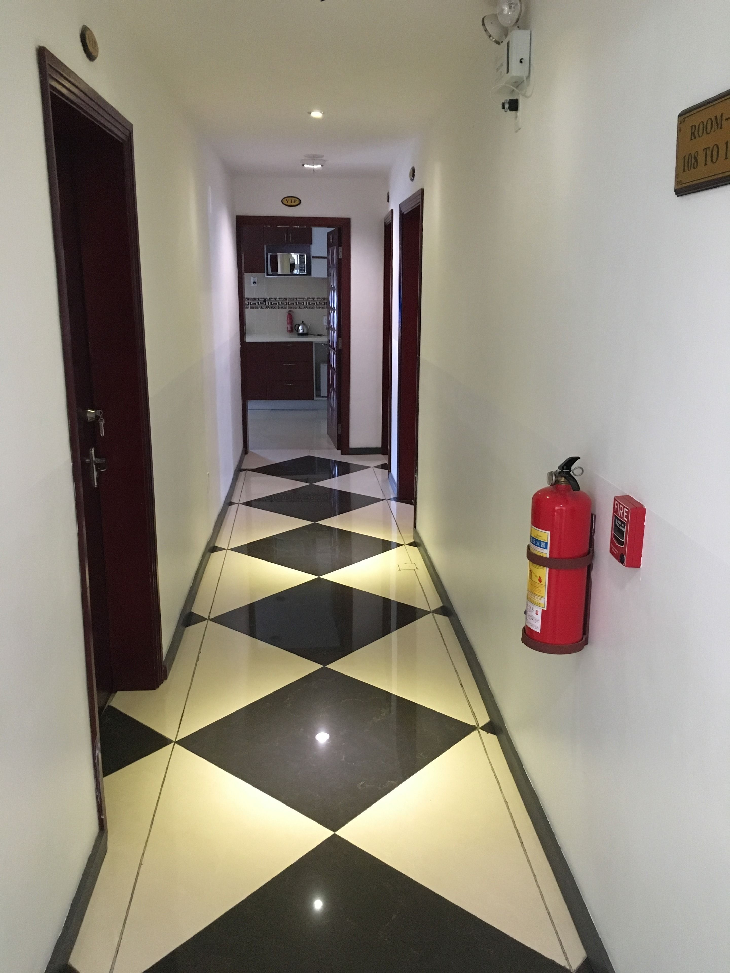 A hotel hallway with a checkered floor, a fire extinguisher mounted on the wall, and a sign indicating room numbers.