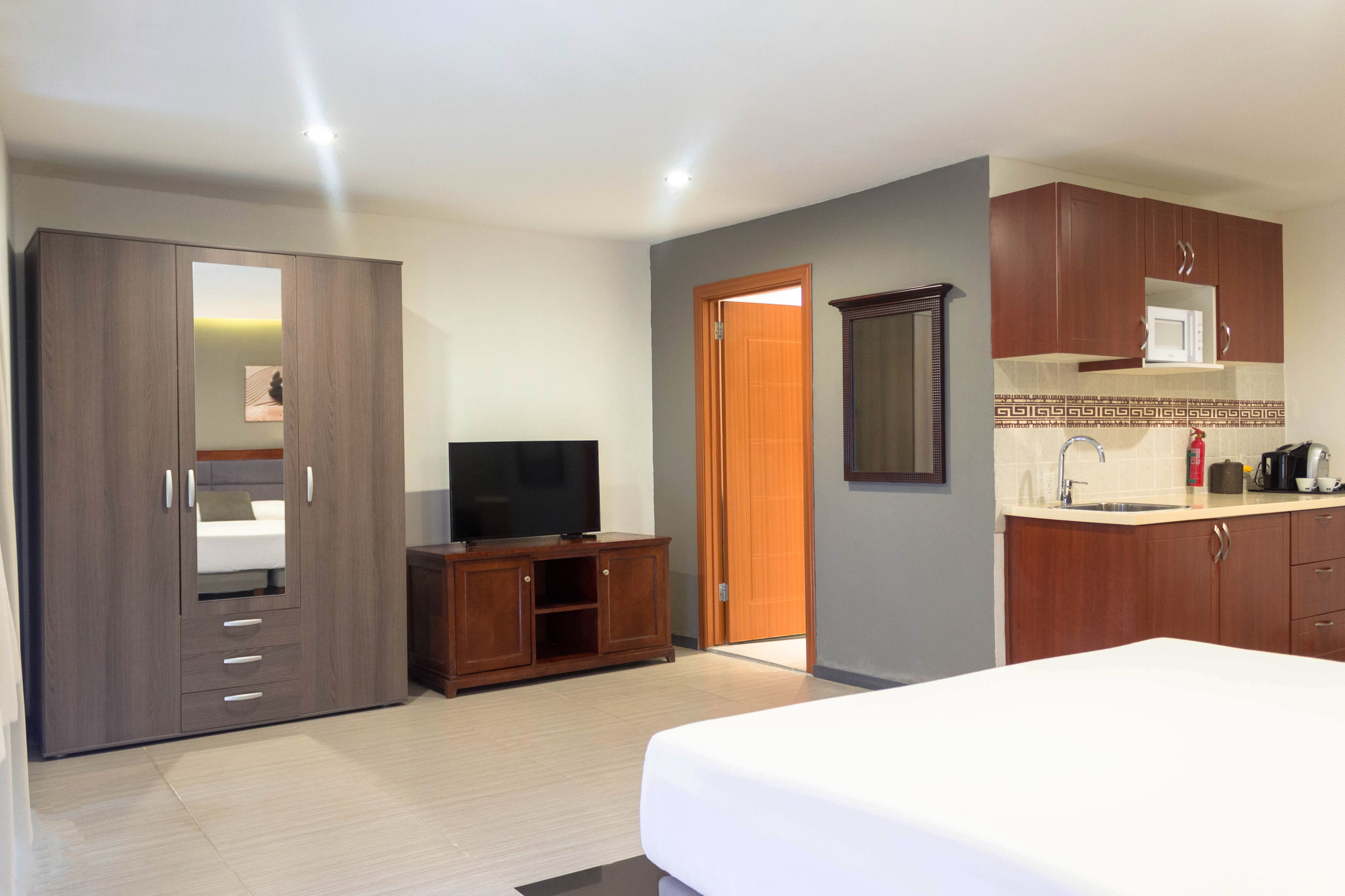 Quarto de hotel com cama, televisão, guarda-roupa, banheiro e cozinha compacta.
