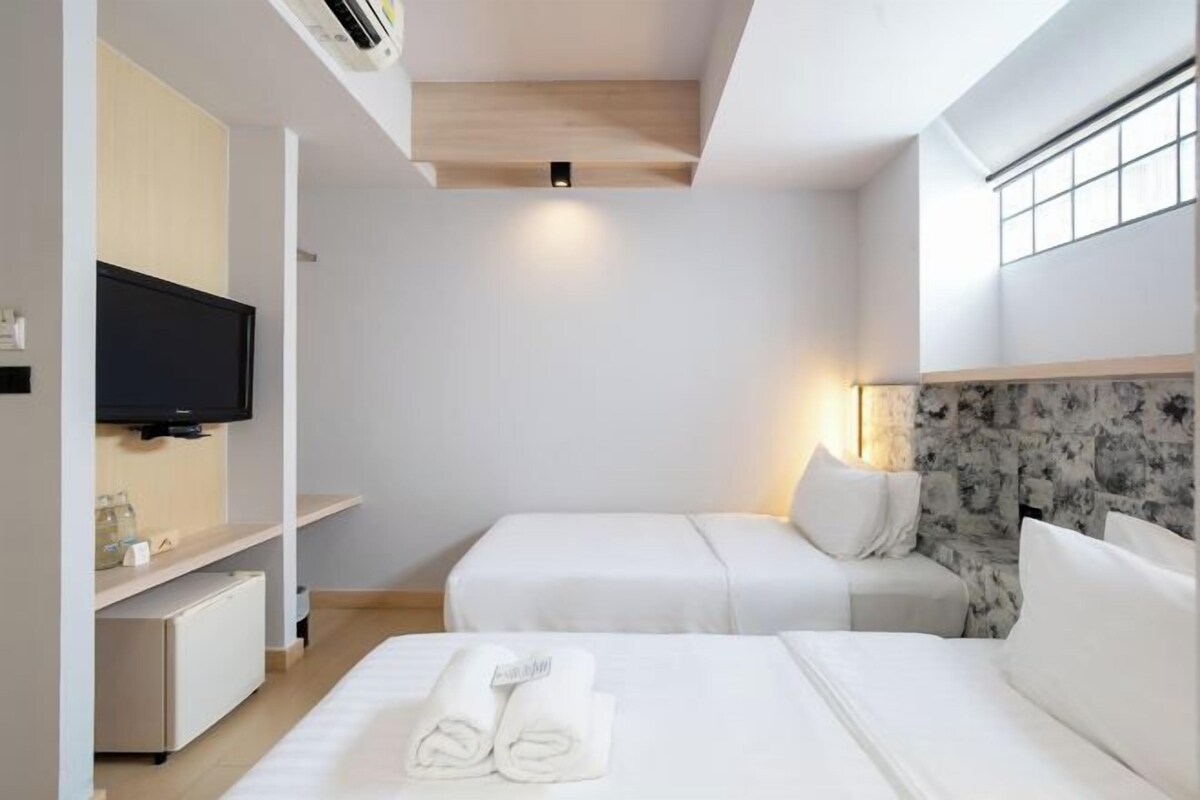 Habitación de hotel con dos camas, un televisor de pantalla plana, una pared decorativa de mármol y un pequeño escritorio con lámpara.