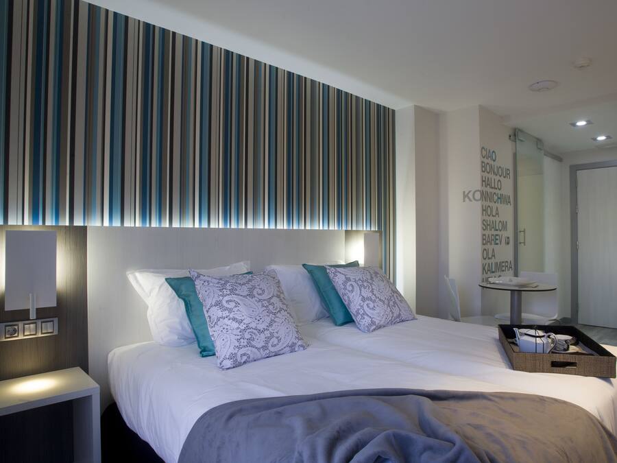 Et moderne hotelværelse med en stor seng, to puder, et natbord med lampe, og et lyspanel på væggen.