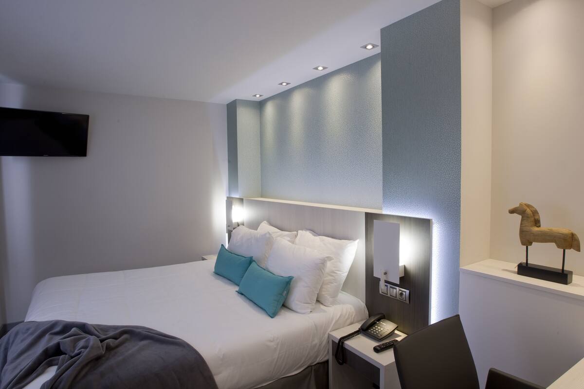 Een moderne hotelkamer met een net opgemaakt bed, een nachtkastje met telefoon en afstandsbediening, en een wandtelevisie.