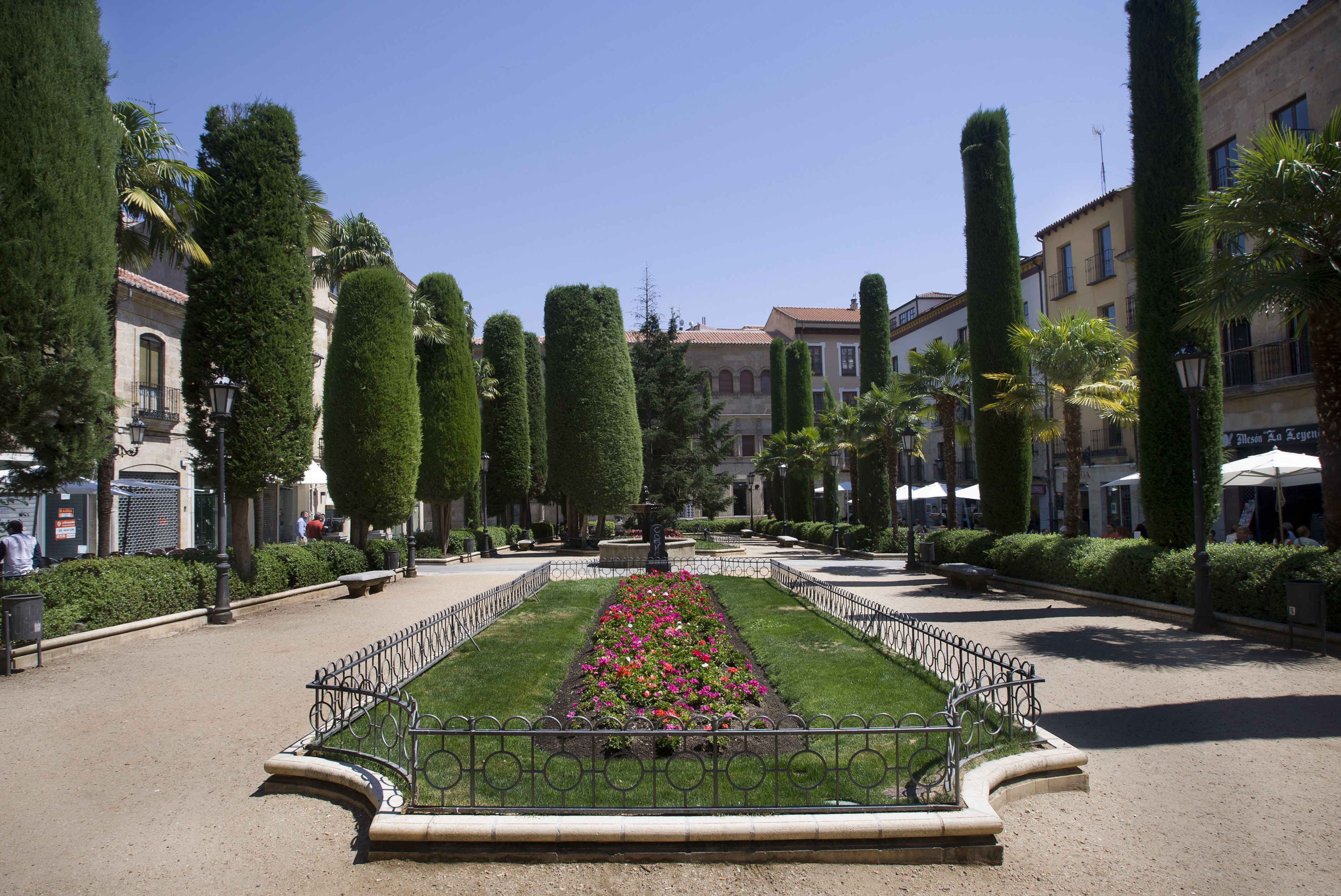 A formal garden with a central flower bed, trimmed hedges, and a paved walkway.