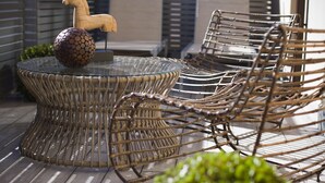 Een ronde glazen tafel met een stoel van geweven materiaal, een decoratief houten paard en een bolvormig object met een bijzondere textuur.