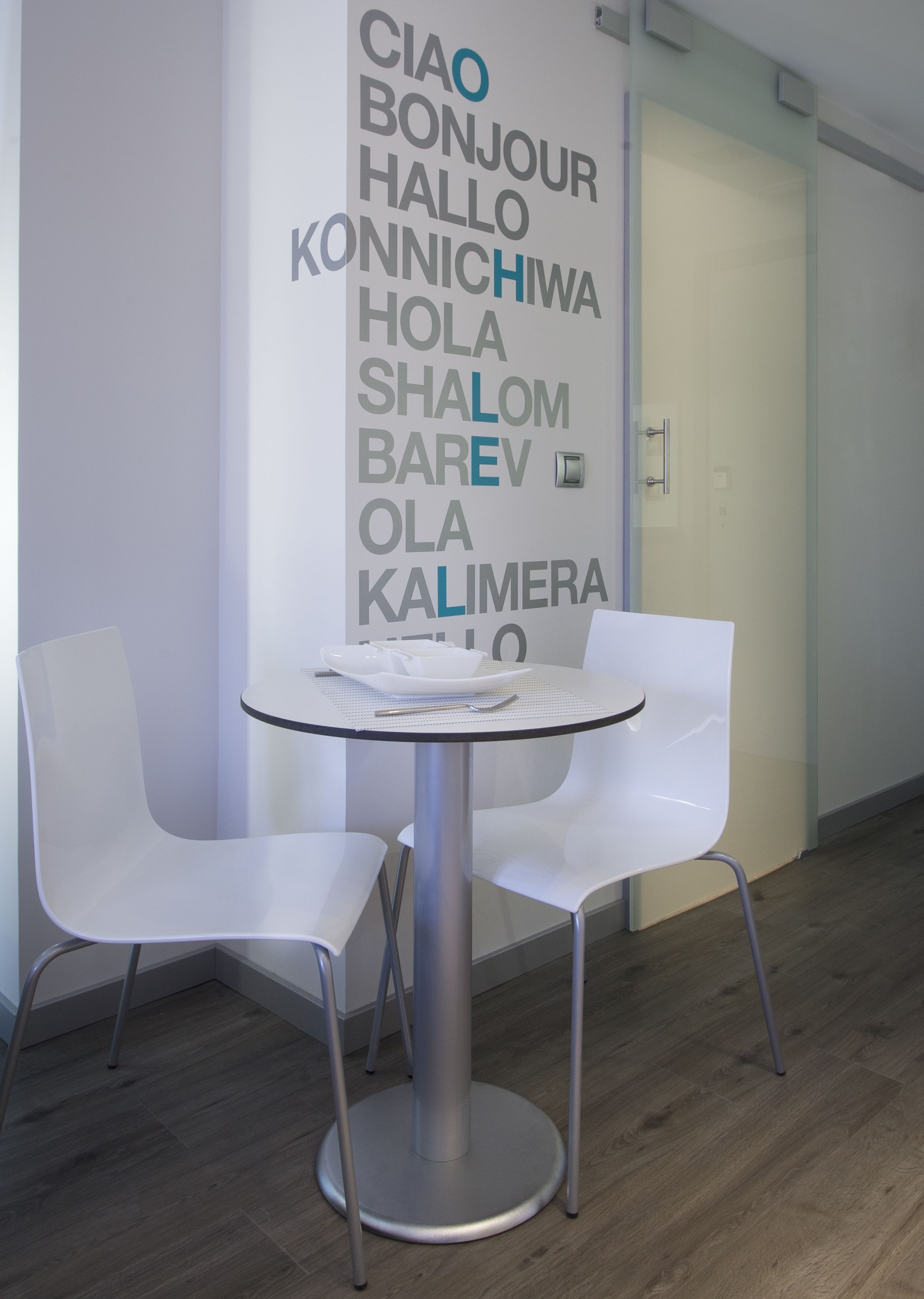 A modern office space with a round table and two white chairs. A wall-mounted sign displays multilingual greetings.