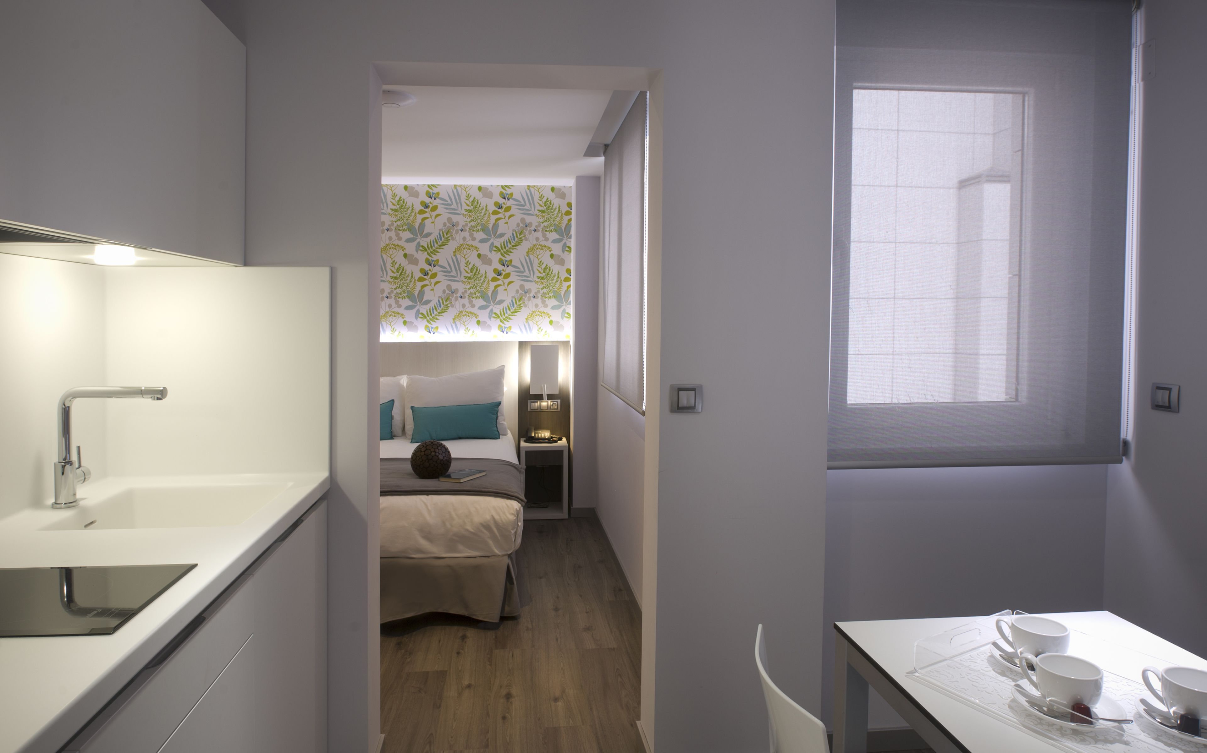 Una habitación de hotel pequeña y funcional con kitchenette, una cama individual, una mesita de noche con lámpara y una ventana con vistas.