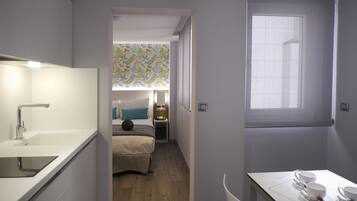 A compact hotel room with a kitchenette, a small bed, a bedside table with a lamp, and a window with a view.