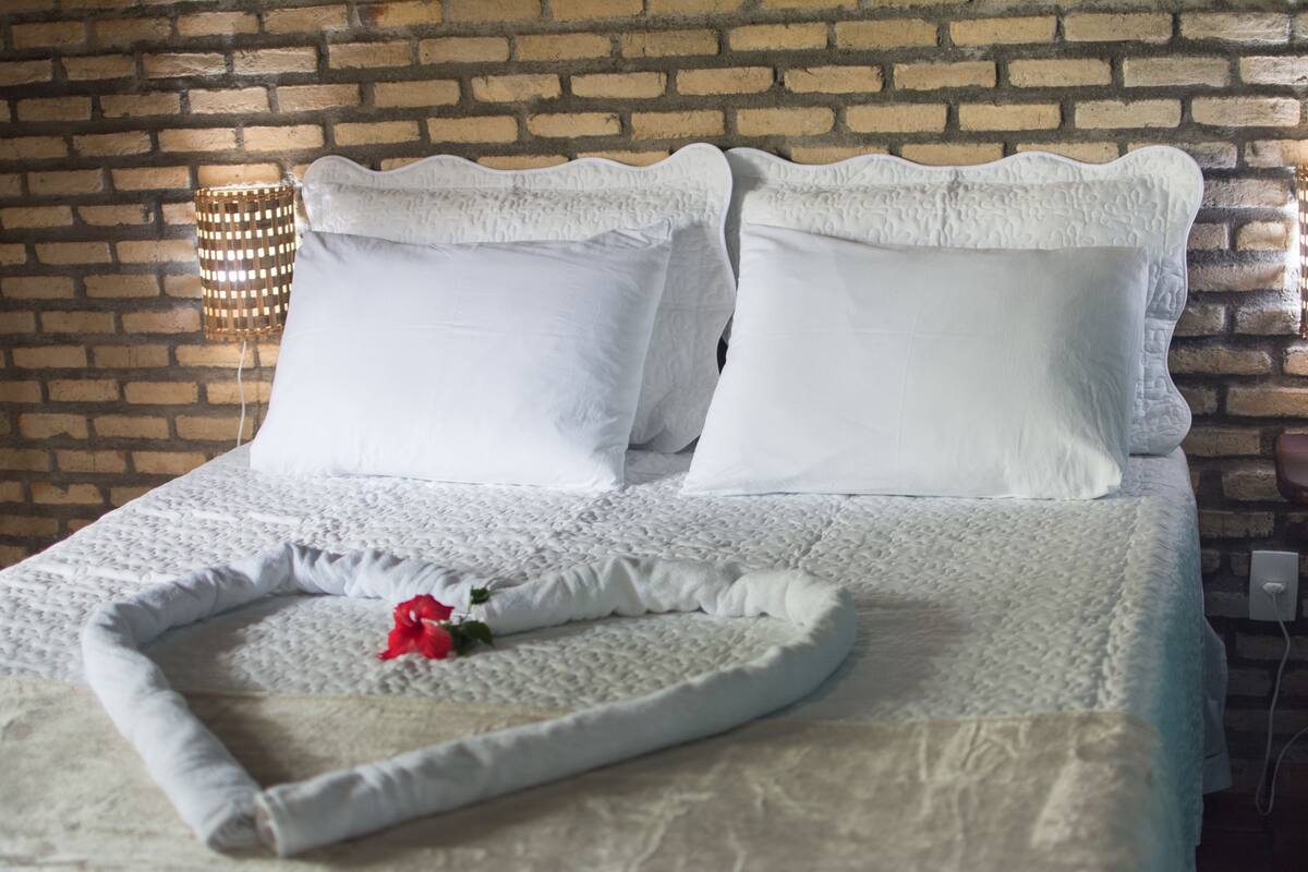 A bed with a white quilt and pillows, a red rose, and a brick wall with a woven light fixture.