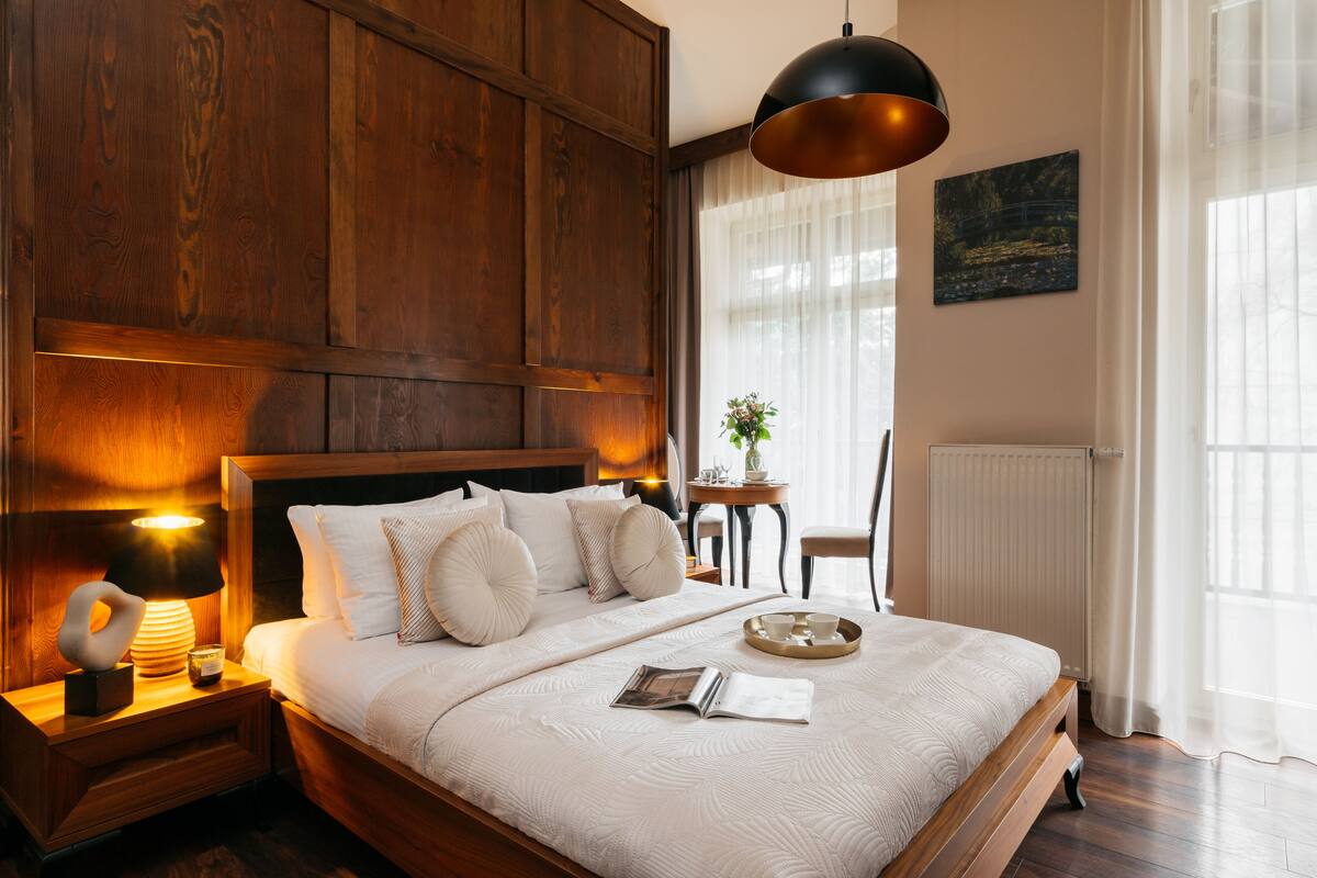 A neatly made bed with a headboard, a bedside table with a lamp, a small table with a plant, and a chair.