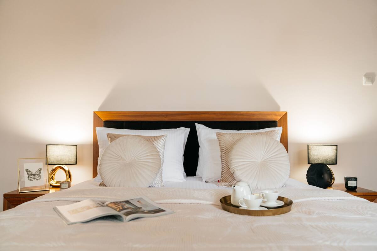 A neatly made bed with white bedding, two bedside lamps, a tray with cups and a teapot, and a framed picture on the nightstand.