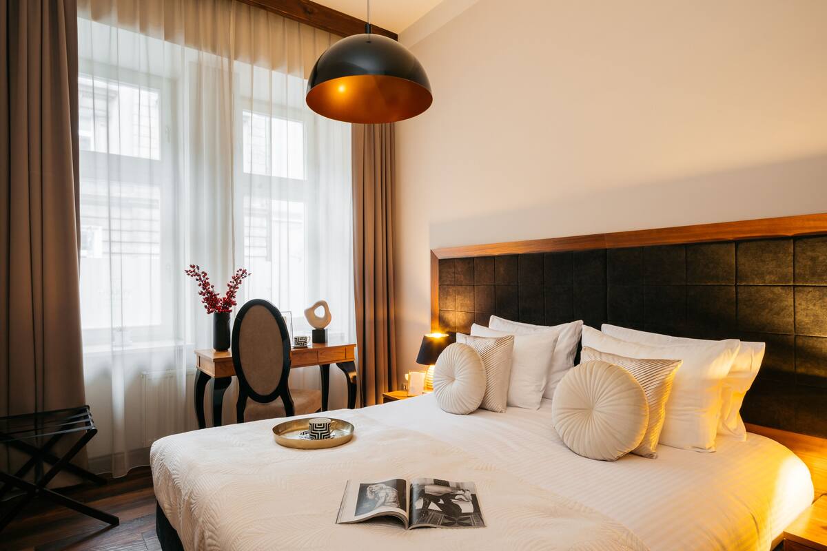A neatly arranged hotel room with a large bed, a bedside table, a reading lamp, and a view of the outdoors through sheer curtains.