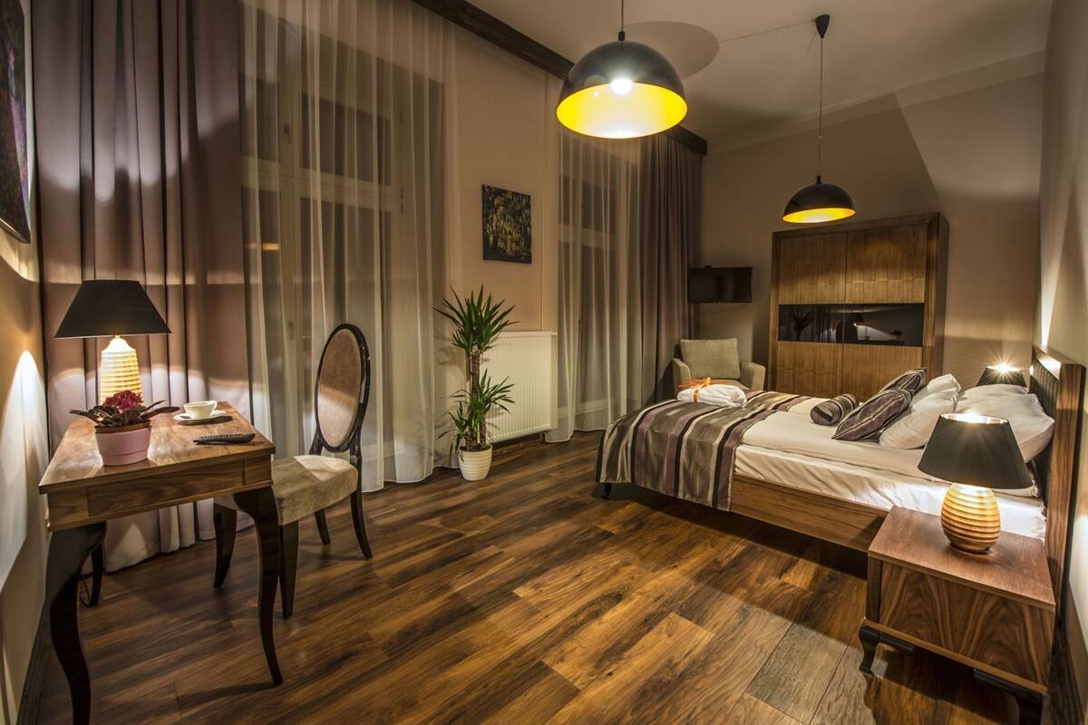 A hotel room with a bed, bedside table, lamp, chair, desk, and a potted plant.