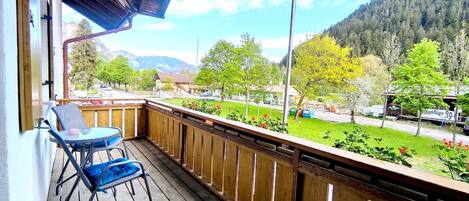 Un balcon en bois, agrémenté d’une table et de chaises, qui offre une vue sur un paysage verdoyant, avec des arbres et une montagne en arrière-plan.