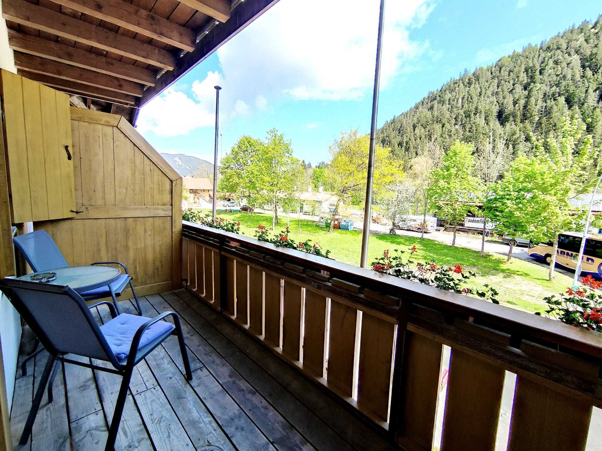 Ein Holzbalkon mit Tisch und Stühlen, der auf eine grüne Landschaft mit Bäumen und einem Berg im Hintergrund blickt.