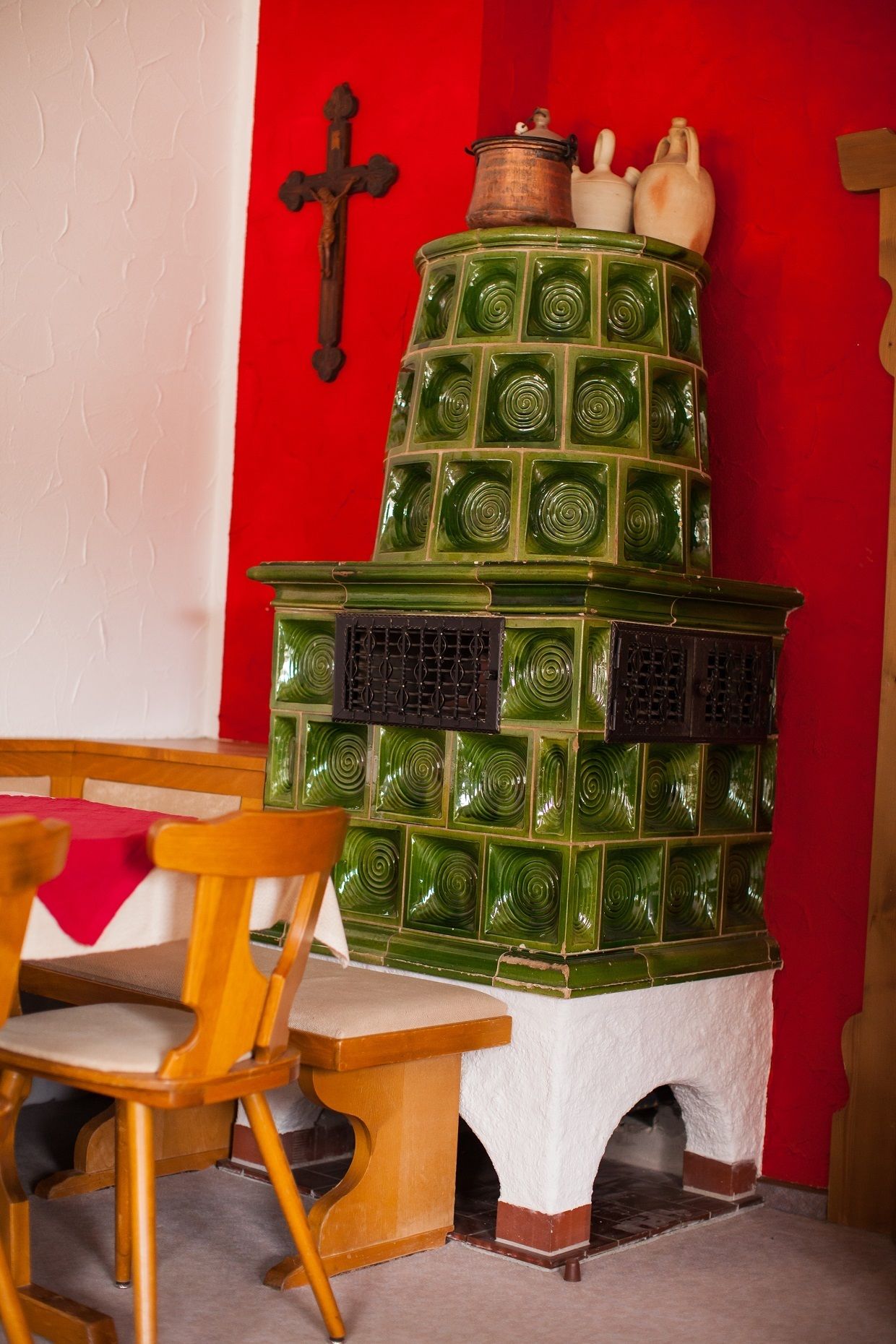 A green stove with a white base, a wooden chair, and a red wall with a cross.
