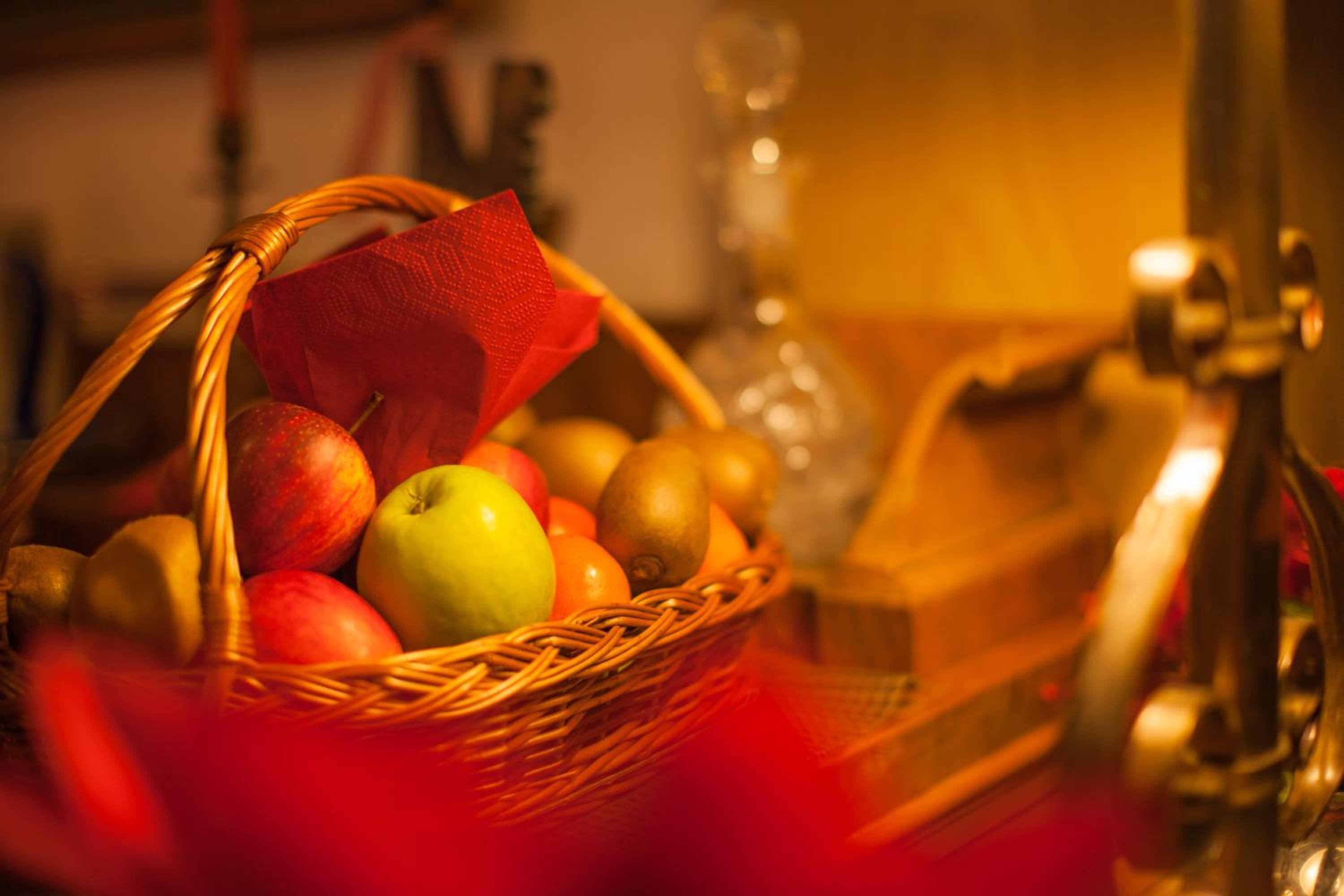 A wicker basket filled with various fruits, including apples and oranges, placed on a surface with a red cloth.